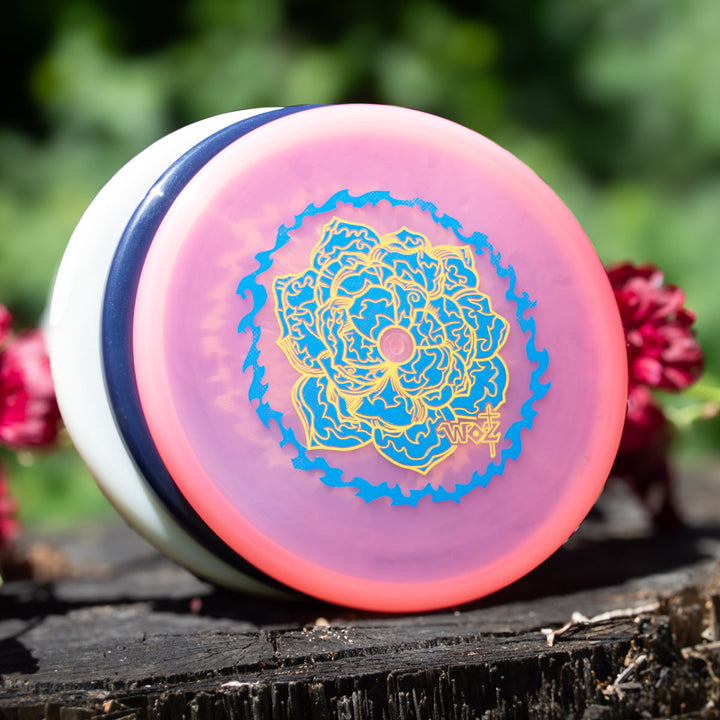 A limited edition WoznBloom Diamond Shaman yo-yo in pink and white with blue and yellow floral patterns sits on a tree stump amid green leaves and red flowers.