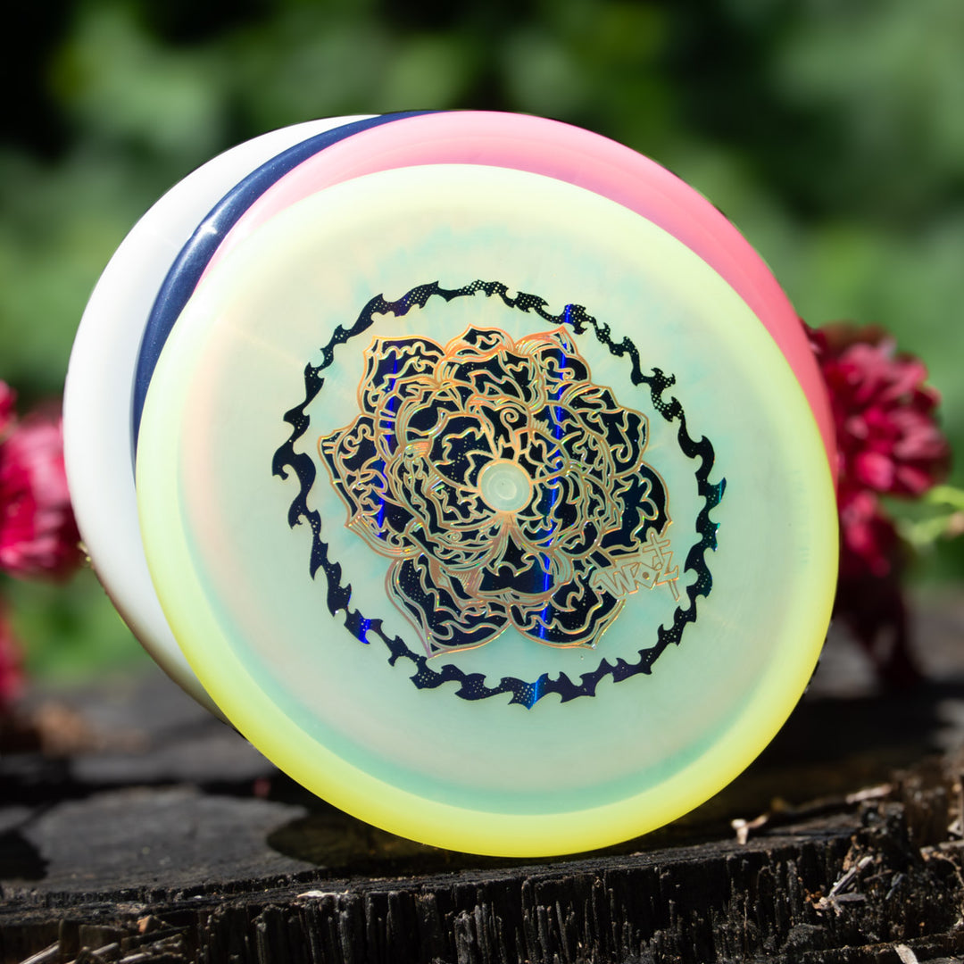 A close-up of three stacked disc golf discs, with the front disc showing the WoznBloom Diamond Shaman special release design, placed on a tree stump outdoors amid blurred greenery and pink flowers in the background.