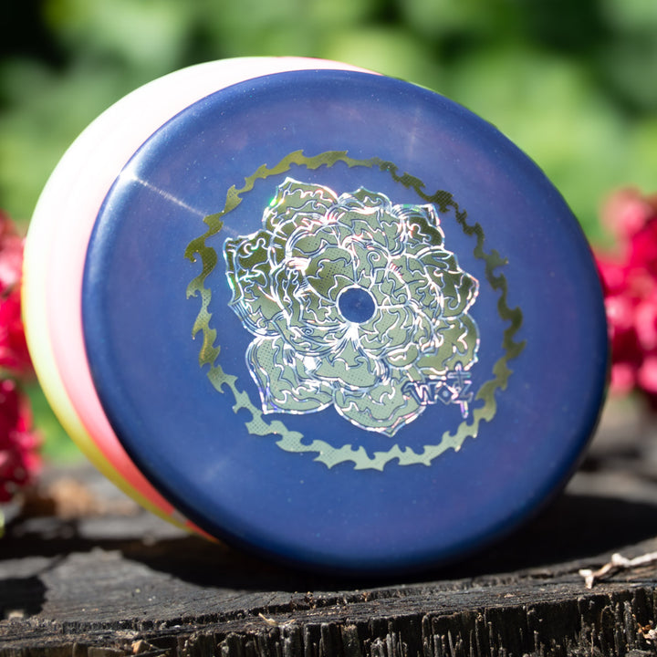 The WoznBloom Diamond Shaman yo-yo, with a blue and pink exterior and detailed floral design, rests on a wooden surface outdoors—a special release ideal for collectors.