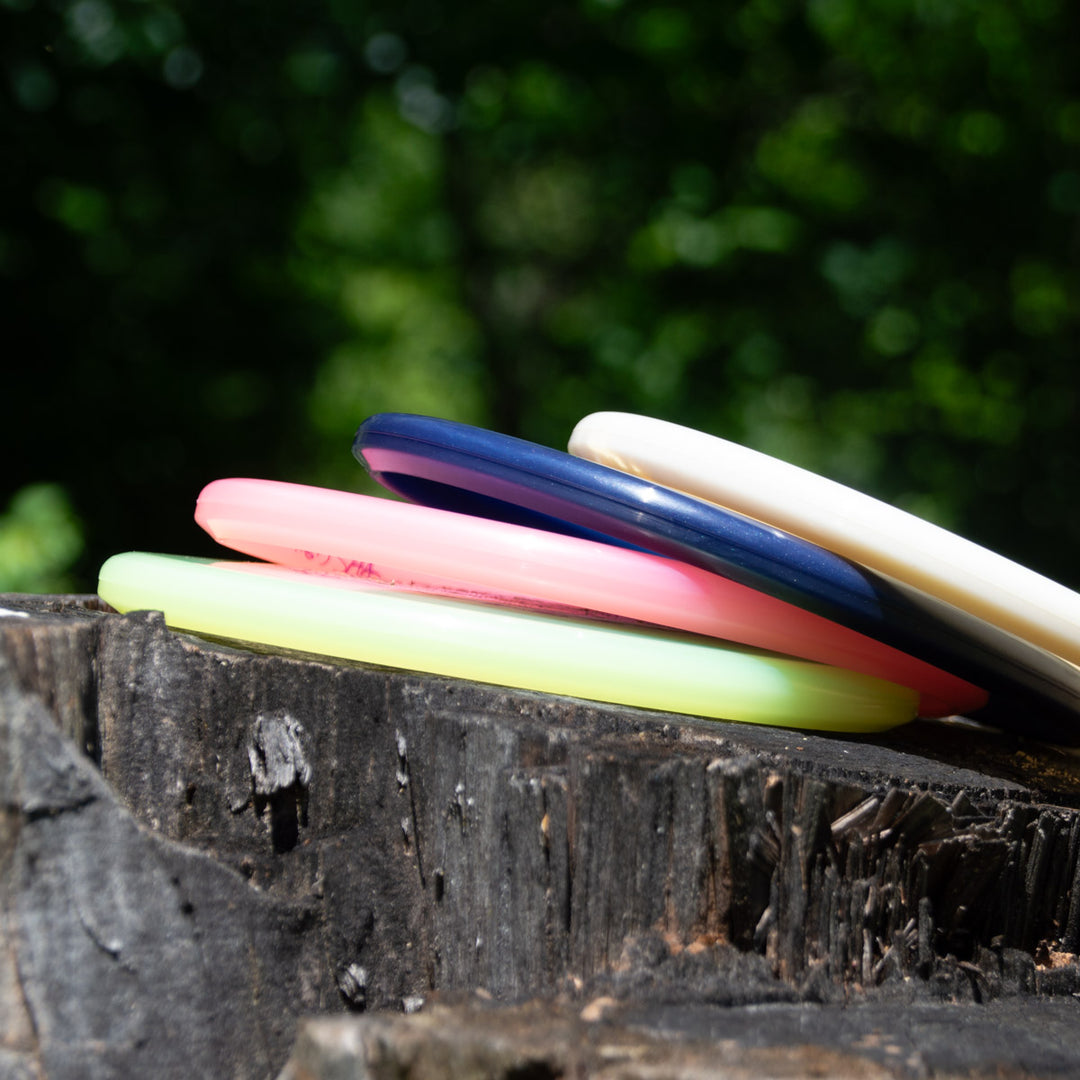 Four colorful disc golf discs, featuring the WoznBloom Diamond Shaman special release, are stacked on a tree stump outdoors with a blurred green forest in the background.