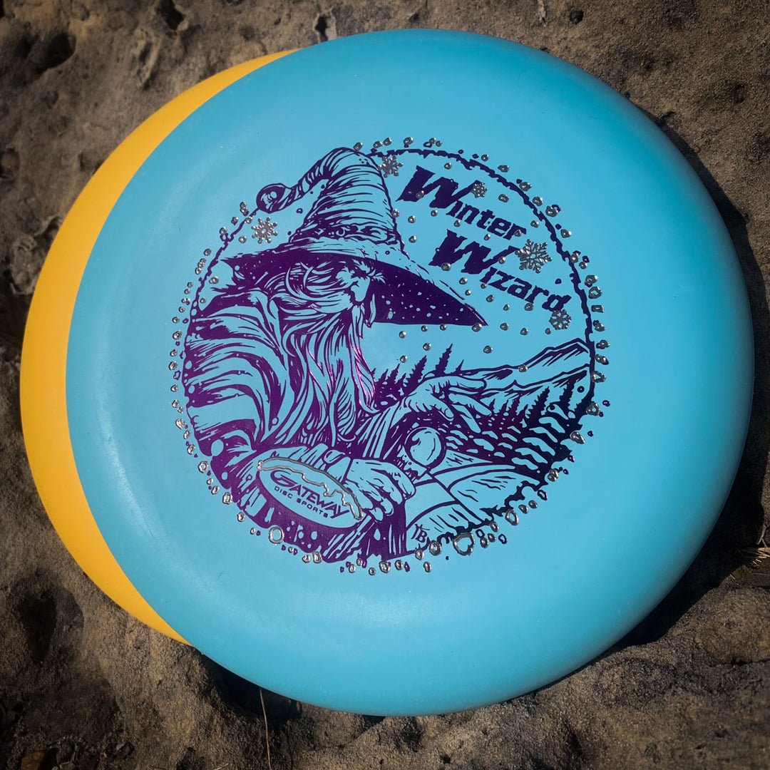 The 2-Disc Players Pack - Winter Wizards 2025 features a blue disc with a "Winter Wizard" design displayed atop a yellow disc on sandy ground.