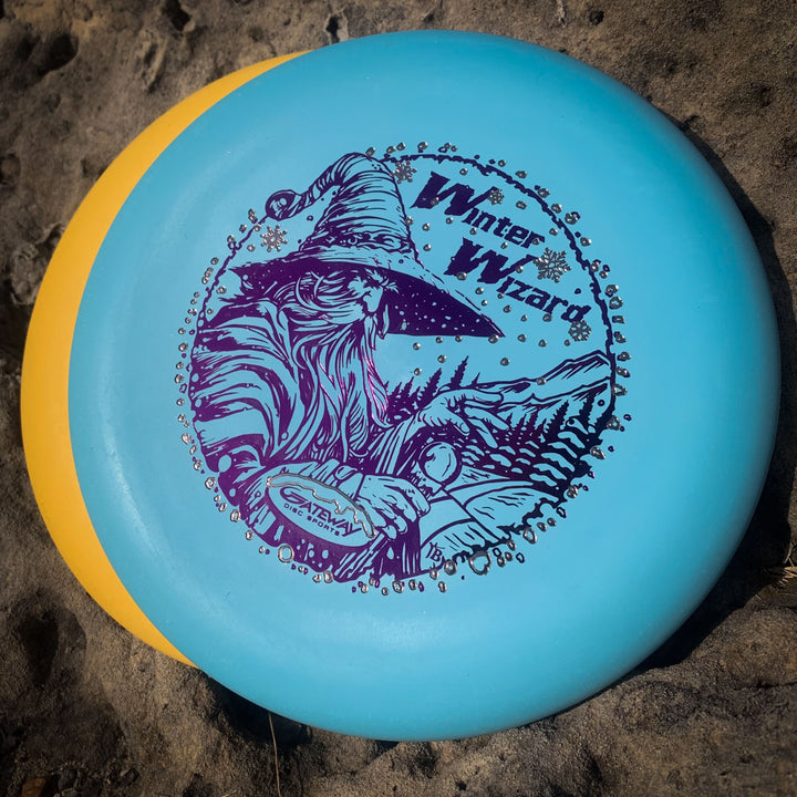 The 2-Disc Players Pack - Winter Wizards 2025 features a blue disc with a "Winter Wizard" design displayed atop a yellow disc on sandy ground.