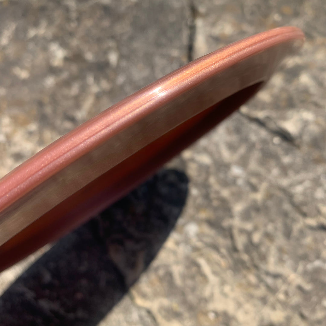 Close-up of the rim of the Apex - Diamond, a reddish-brown high-speed driver disc, viewed at an angle with a rough stone background.