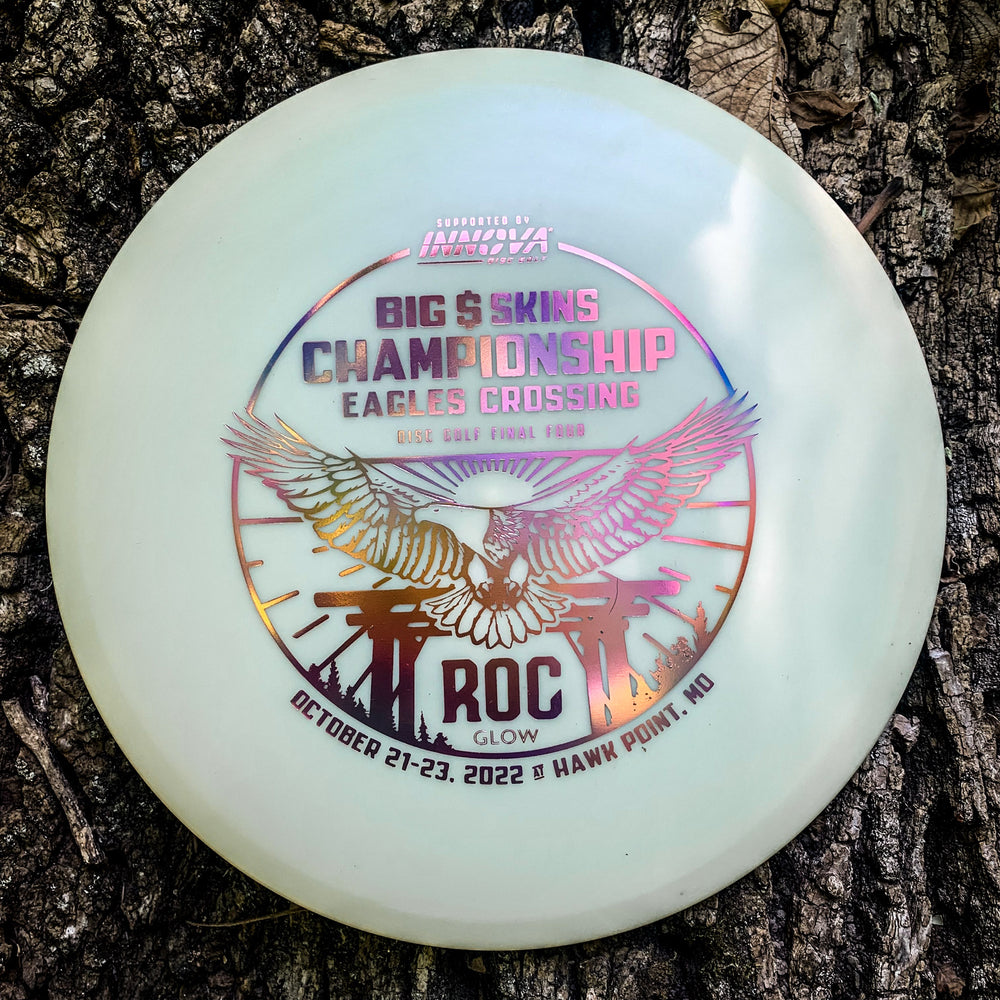 The EC Innova Roc DX Glow disc, featuring "Big & Skins Championship Eagles Crossing ROC Glow," dates October 21-23, 2022, Hawk Point, MO, and honoring the Innova Roc, rests on tree bark.