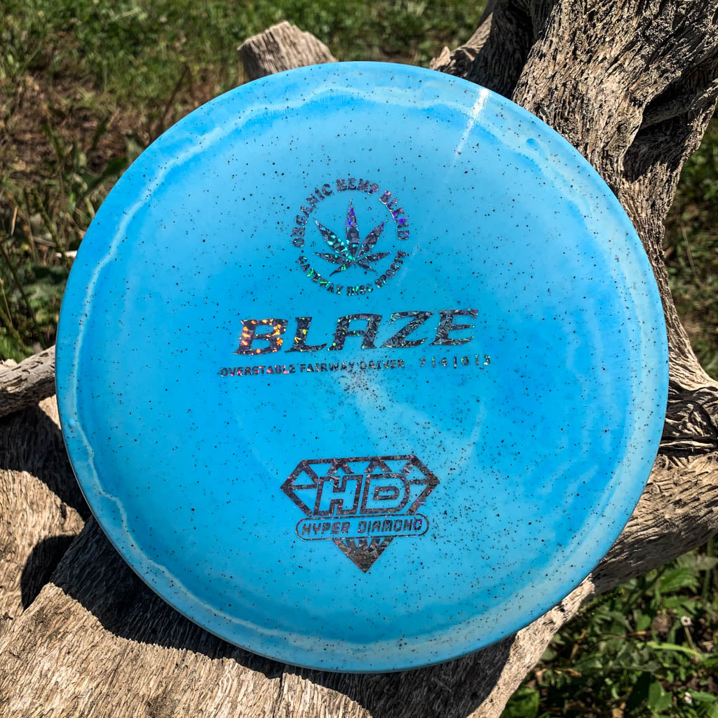 A blue Blaze - Hyper Diamond (HD) Hemp disc golf driver, prized for its overstable flight and reliability in headwinds, lies on weathered wood outdoors with dirt speckling its surface.