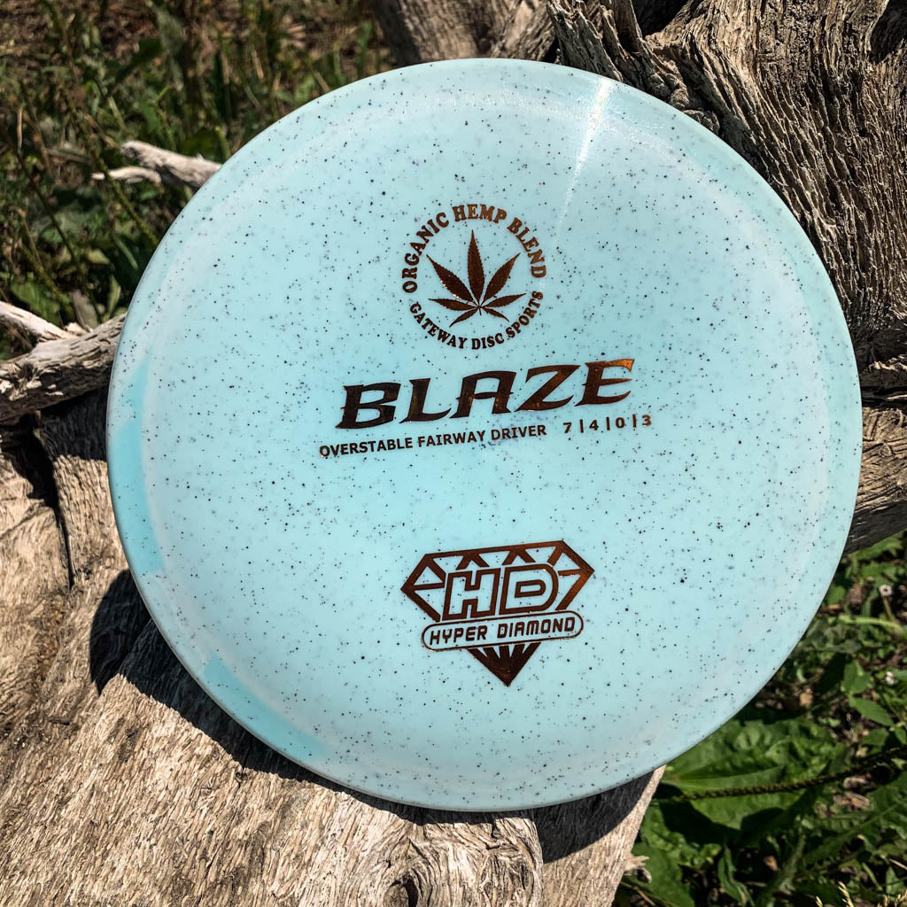 A light blue speckled Blaze - Hyper Diamond (HD) Hemp overstable disc golf fairway driver sits on a weathered tree trunk outdoors, ready to tackle headwinds and deliver a controlled, predictable flight.