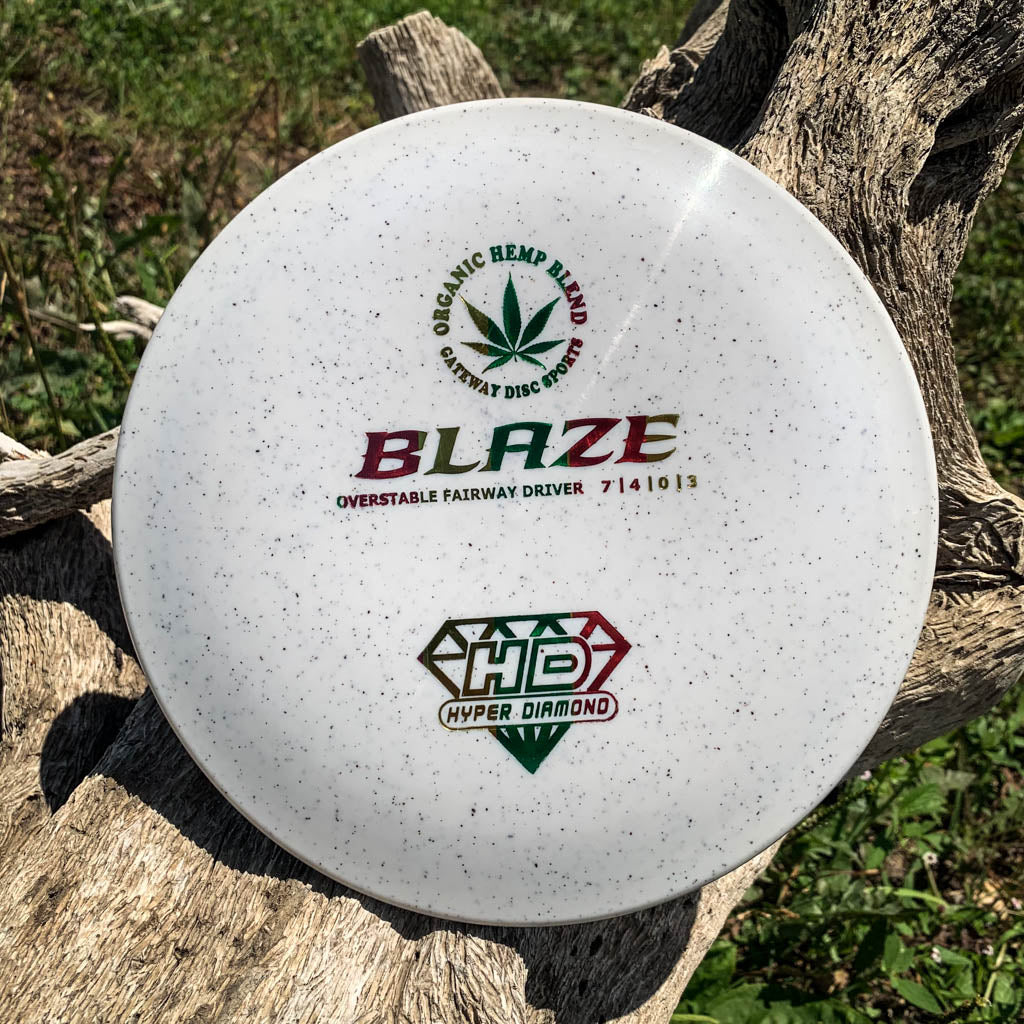 The Blaze - Hyper Diamond (HD) Hemp, a white speckled overstable disc golf driver with a hemp leaf graphic, sits on rough wood outdoors, offering reliable performance and predictable flights, even in headwinds.