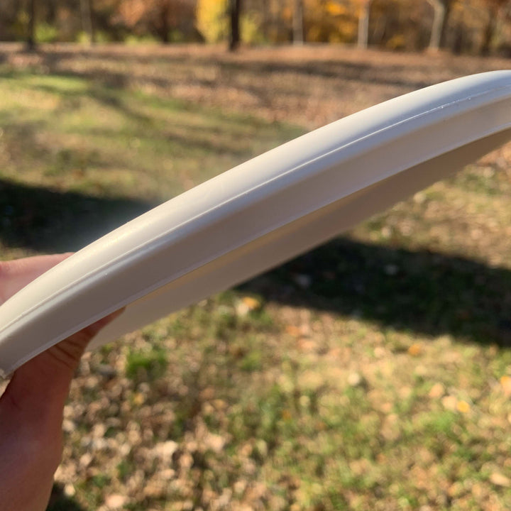 A hand holds a Chief OS - Pure White disc outdoors, with grass and trees in the background.