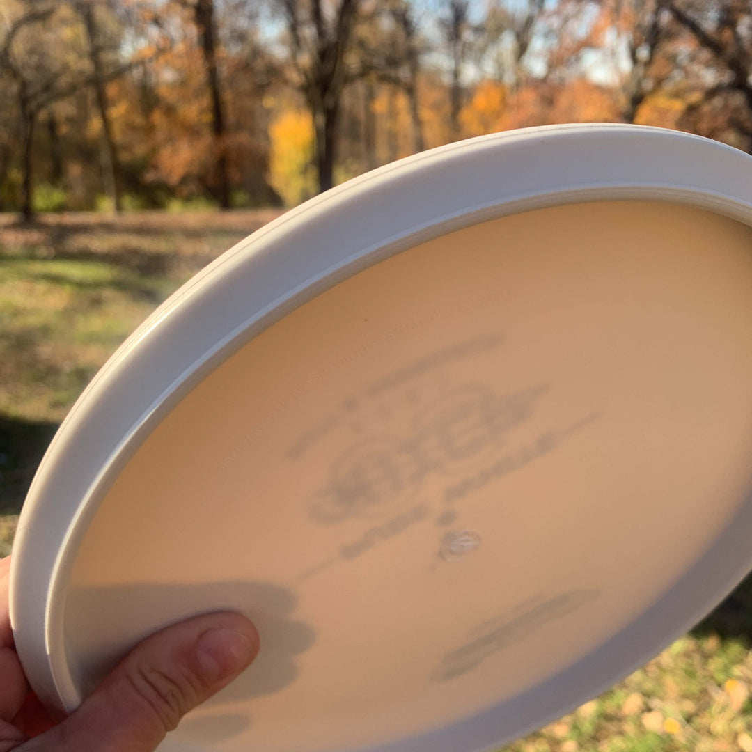 A hand holds a Chief OS - Pure White disc outdoors, made from Premium Suregrip Plastic, with blurred trees and autumn leaves in the background.