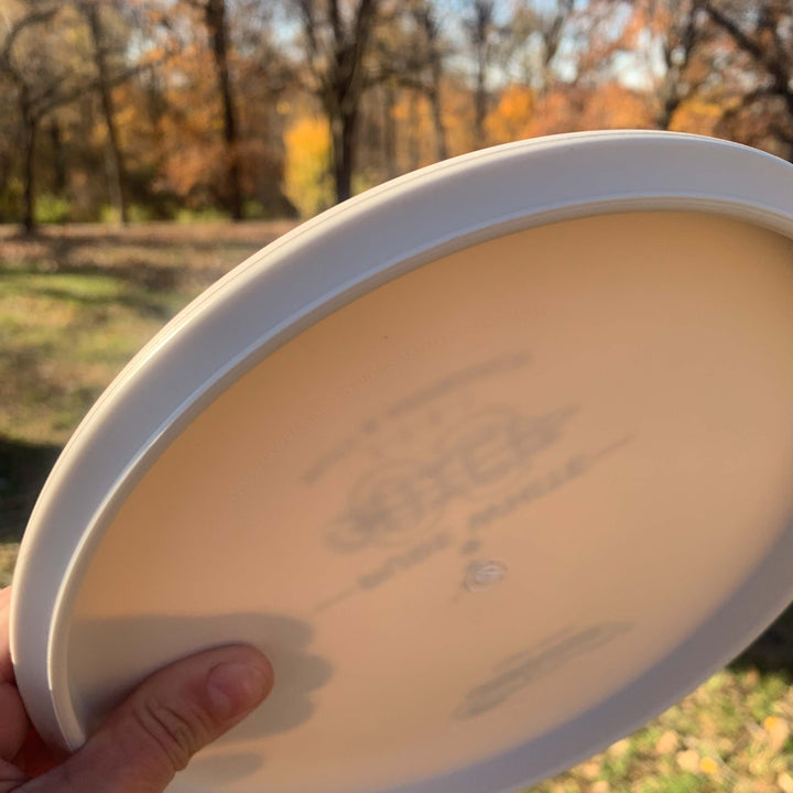 A hand holds a Chief OS - Pure White disc outdoors, made from Premium Suregrip Plastic, with blurred trees and autumn leaves in the background.