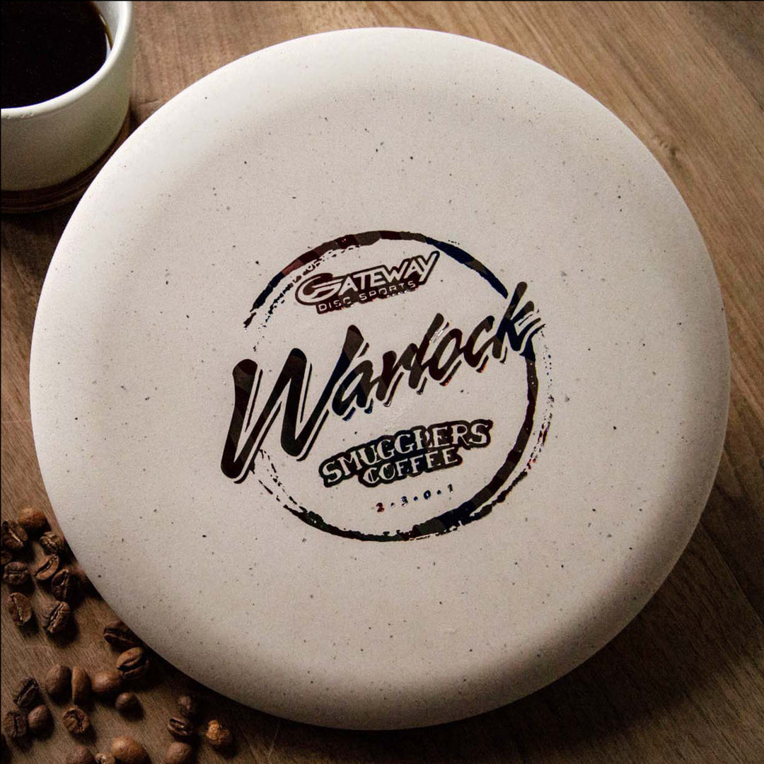 A Warlock - Smugglers Coffee Special Blend disc with "Gateway Disc Sports Warrack Smugglers Coffee" sits on wood beside coffee beans, a cup of coffee, and an espresso blend worthy of the Tomb of Caffeination.