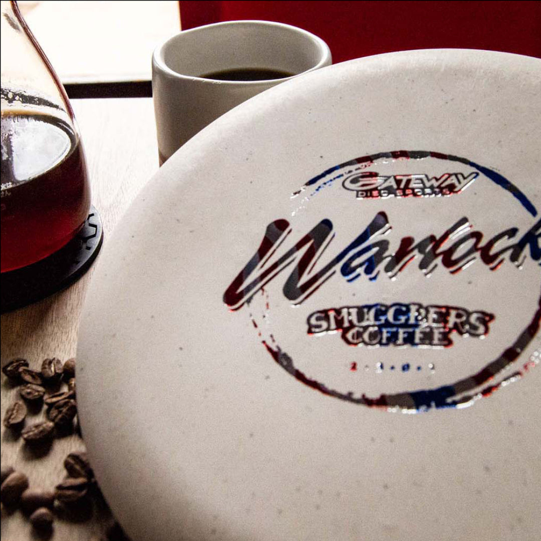 A close-up of the Warlock - Smugglers Coffee Special Blend disc golf disc, featuring "Smugglers Coffee" logos, sits beside a cup, carafe, and scattered beans—a tribute to their bold espresso for conquering the Tomb of Caffeination.