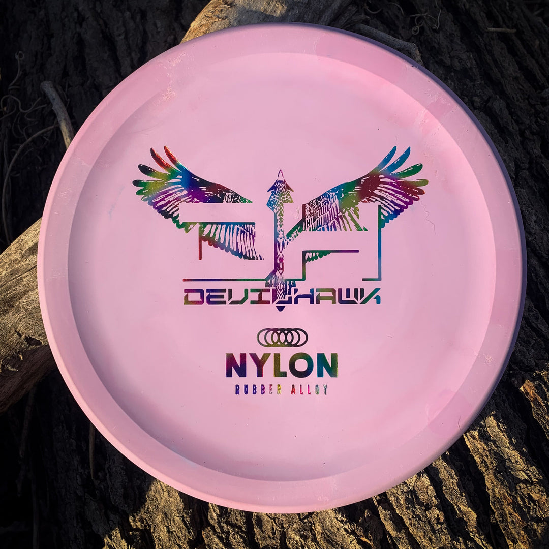 A pink Devilhawk - Suregrip Nylon disc with a colorful winged design, "DEVILHAWK," the Audi logo, and "NYLON RUBBER ALLOY" is a unique over-stable putter with Devil Hawk's signature Thumb Track, shown on rough wood.