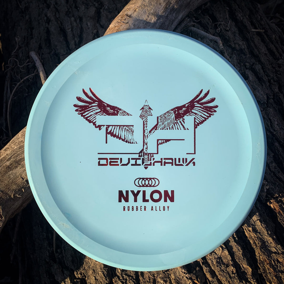 A light blue Devilhawk - Suregrip Nylon disc golf disc with a stylized hawk graphic, Thumb Track design, and "DEVILHAWK" plus "NYLON RUBBER ALLOY" text, resting on tree bark.