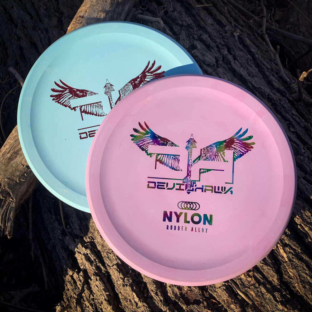 Two Devilhawk - Suregrip Nylon disc golf discs, one light blue and one pink with colorful designs, featuring the unique Thumb Track, rest on dark textured tree bark.