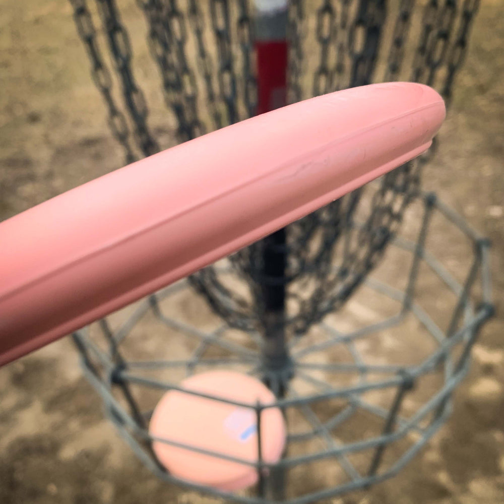 Close-up of the Wizard - Eraser, a pink disc golf disc ideal for mid-range drives, about to land in a metal basket with chains.
