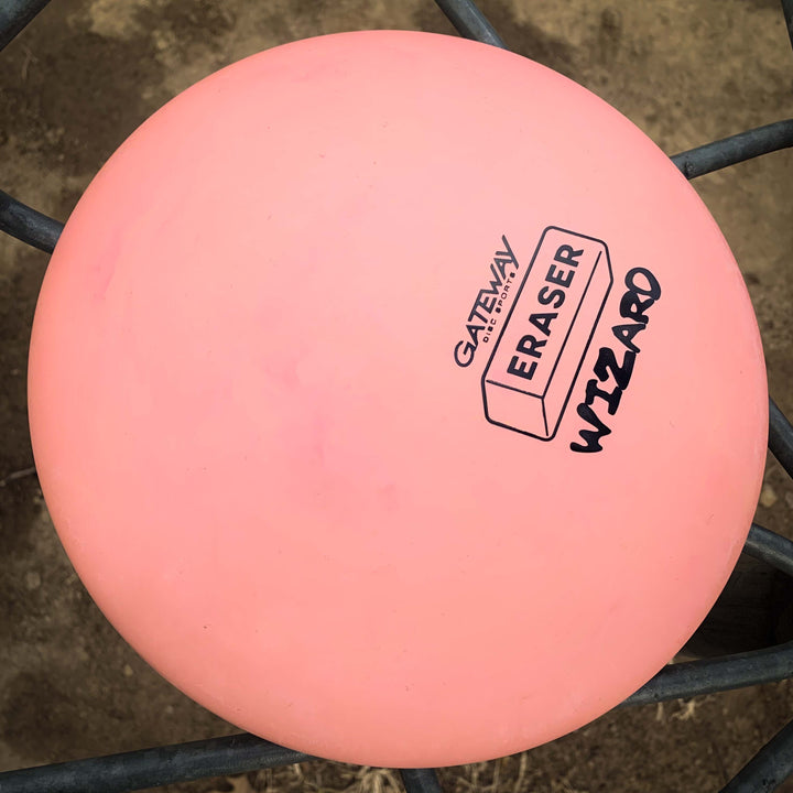 A pink Wizard - Eraser putter by Gateway, famed for its stable flight and reliability, sits on a metal fence. The black brand and model print highlight its suitability for confident putts and mid-range drives, even in windy weather.