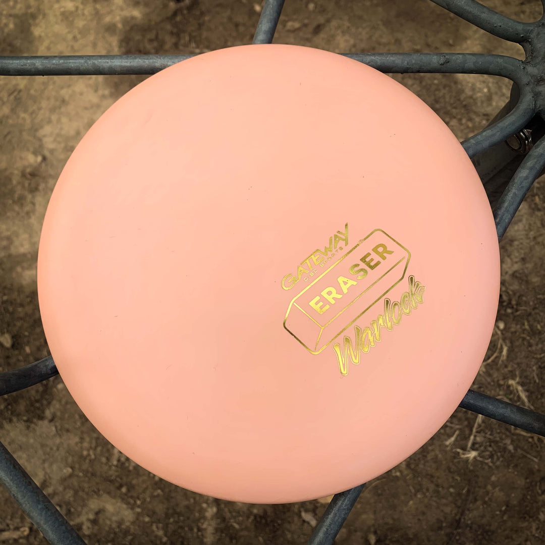A peach-colored Warlock - Eraser disc golf putter with gold "Gateway Eraser" and "Warlock" text sits on a metal bench€”ideal for players wanting a stable putter.