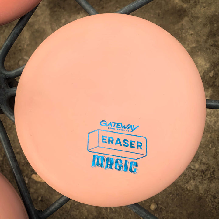 A pink, beginner-friendly Magic - Eraser disc golf putter with blue "Gateway Disc Sports" text rests on a metal rack above dirt.