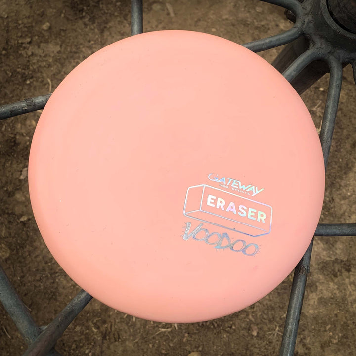A Voodoo - Eraser putter in pink is placed on a metal surface, with dirt visible in the background.
