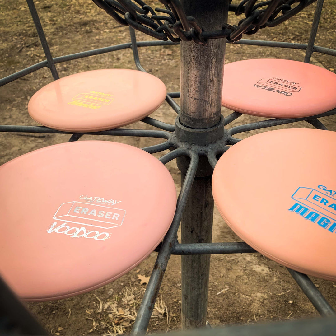 Several pink Voodoo - Eraser putter discs are stacked on the basket of a disc golf target, with metal chains and dry grass visible in the background.