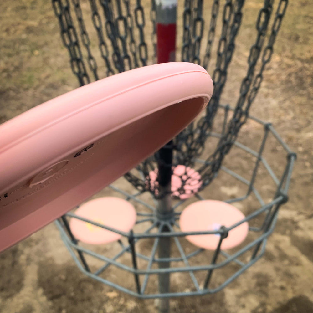 A pink Voodoo - Eraser putter is about to be thrown into a metal disc golf basket on a dirt-covered outdoor course.