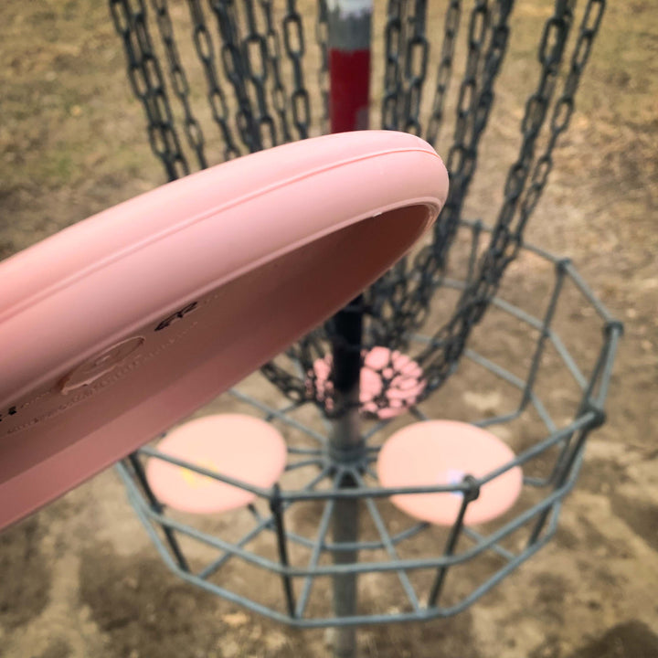 A pink Voodoo - Eraser putter is about to be thrown into a metal disc golf basket on a dirt-covered outdoor course.