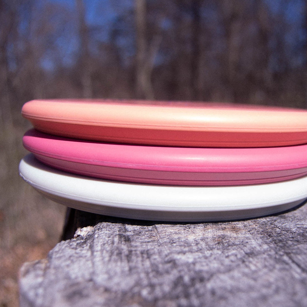 Three Houdini - Eraser discs in peach, pink, and white sit stacked on a tree stump outside, ready for your power-grip backhands or as trusty putt and approach discs—set against a blurred forest background.