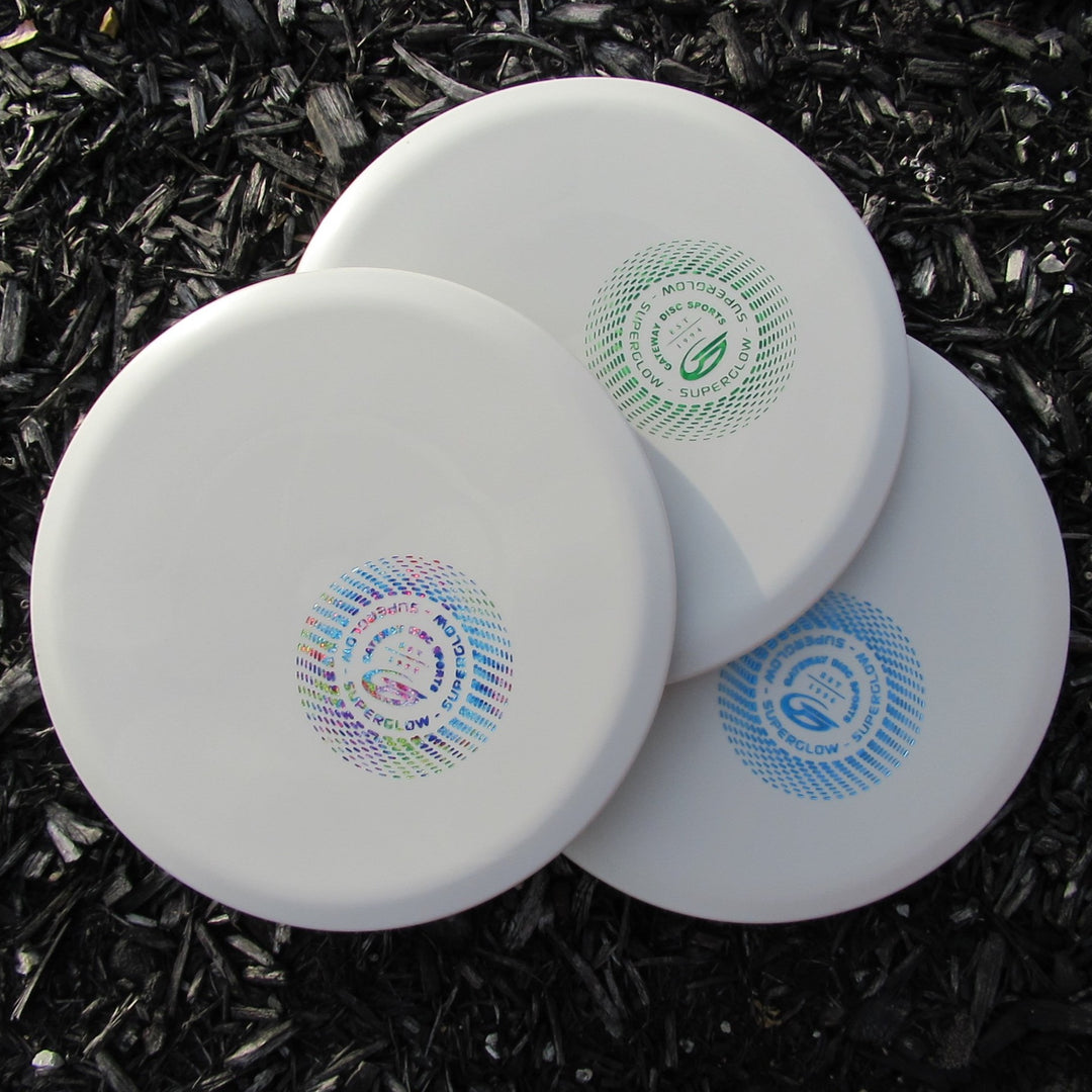 Three white Mystic - SureGrip™ Superglow discs with colorful circular logos overlap on a dark wood chip ground, ideal for players seeking controllable mid-range performance, easy straight flight, and impressive glide.
