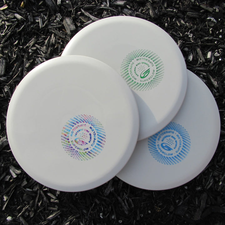 Three white Mystic - SureGrip™ Superglow discs with colorful circular logos overlap on a dark wood chip ground, ideal for players seeking controllable mid-range performance, easy straight flight, and impressive glide.