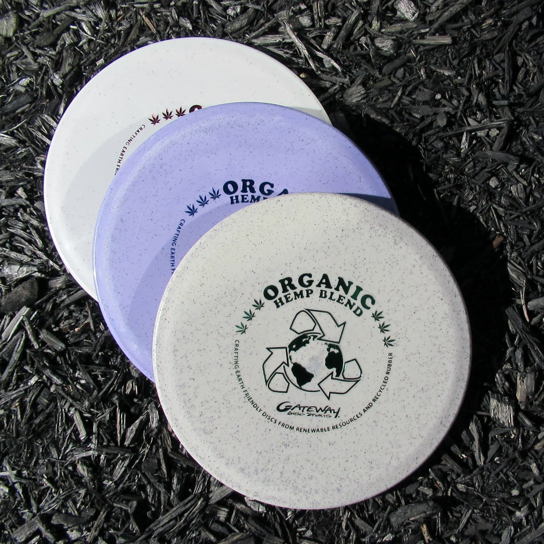 Three Warrior - Organic Hemp mid-range discs are stacked on dark wood chips; the top disc displays a recycling symbol and earth graphic. These overstable discs offer controlled, reliable throws, ideal for precision shots.