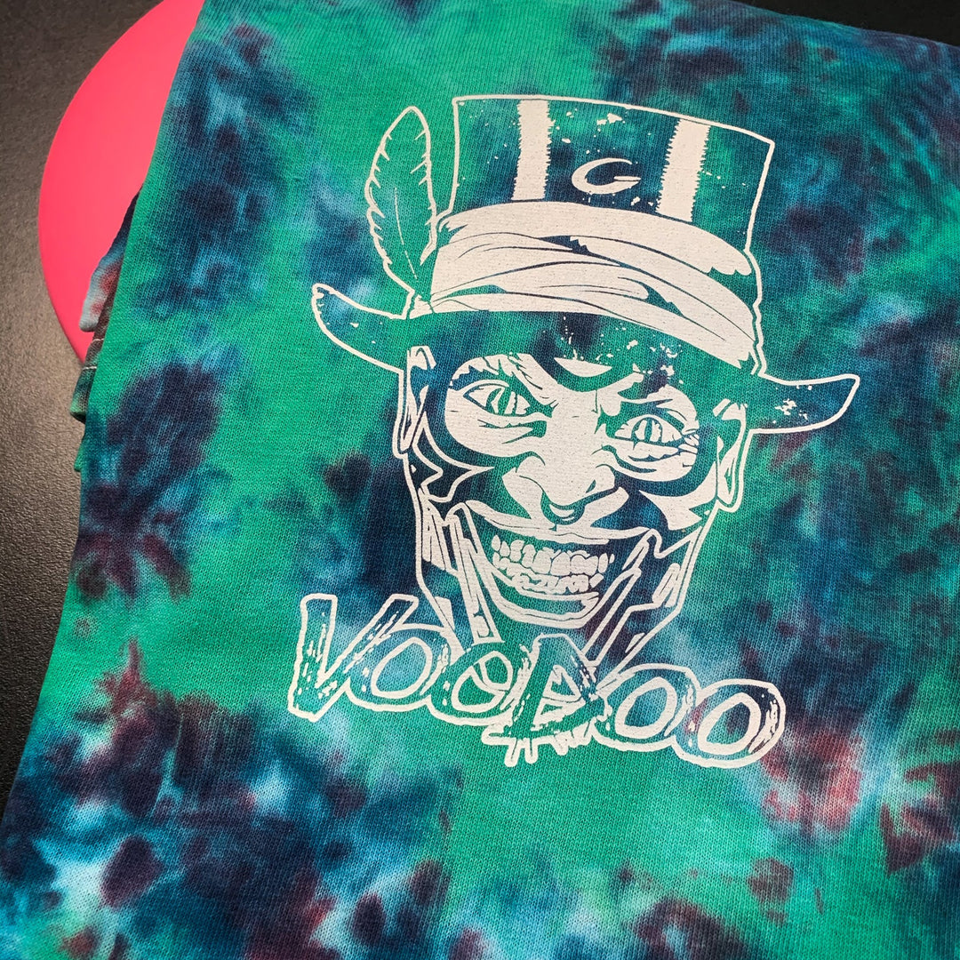 The Tie Die VooDoo Short Sleeve T-shirt is a green and blue tie-dye heavy cotton tee with a white graphic of a smiling face in a top hat and feather, "VOODOO" text below. Machine washable for easy care.
