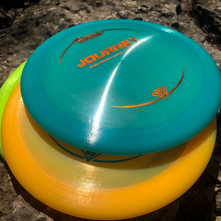 Three brightly colored disc golf discs, the top teal one labeled "Journey - Hyper Diamond (HD)," stacked on a sunlit rock—ideal as a long-range distance driver for players seeking controllable flight and maximum throw distance.