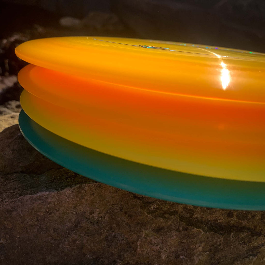 A close-up of four Journey - Hyper Diamond (HD) discs—renowned for controllable flight and long-range distance—stacked in yellow, orange, and blue on a rough rock surface.