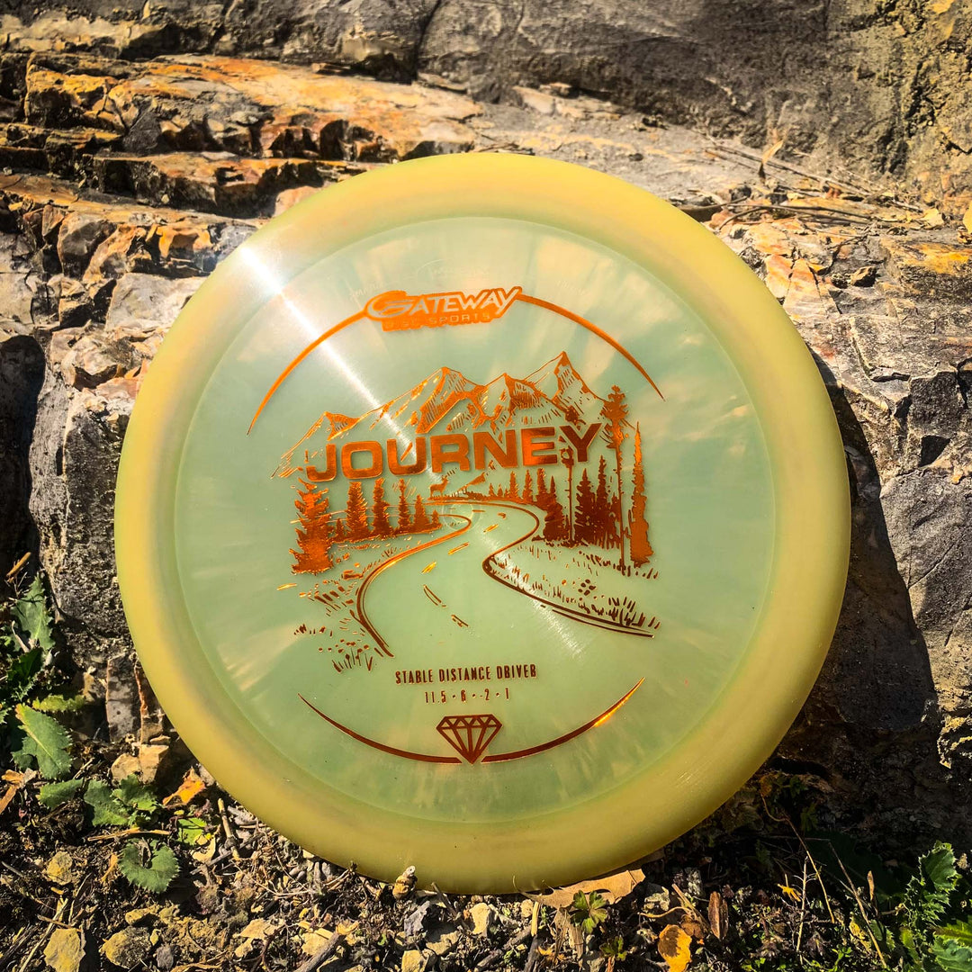 A yellow Gateway Journey - Diamond disc golf disc, celebrated for its controllable flight, rests on a rocky outdoor surface and displays a graphic of mountains with a road.