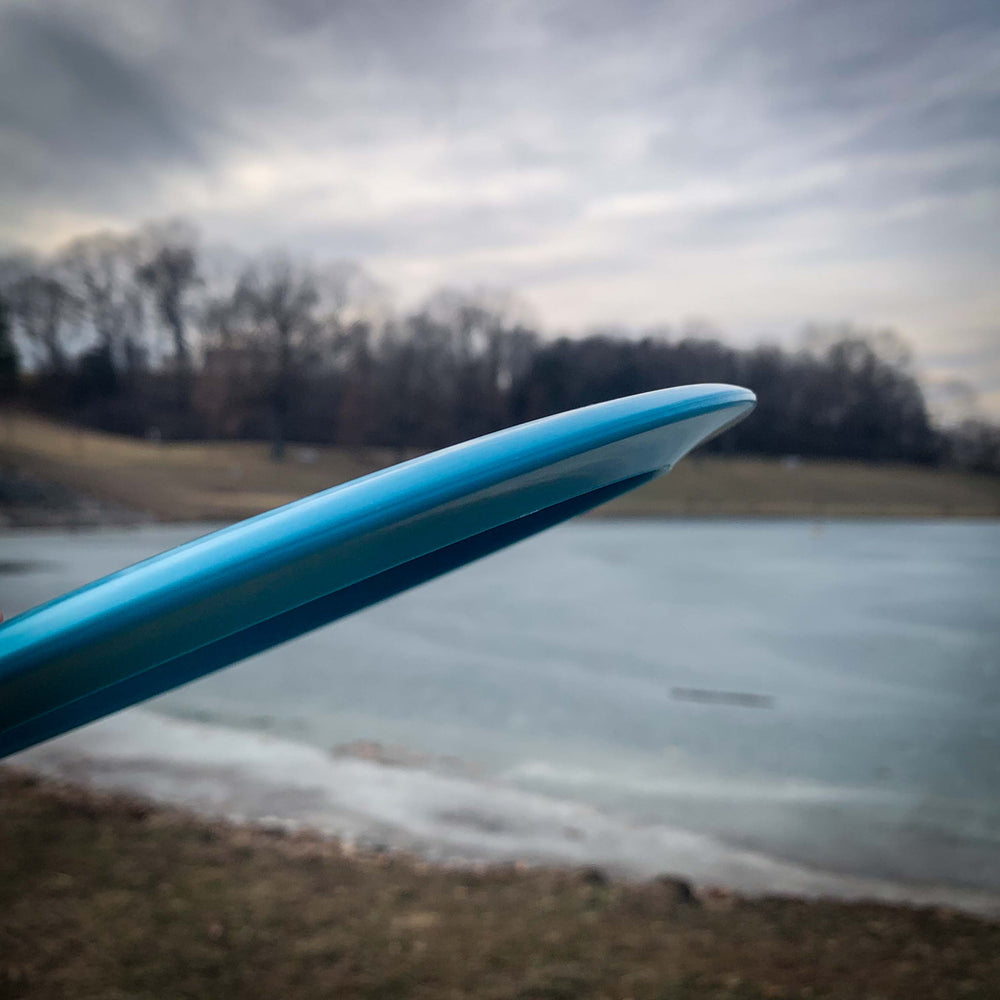 A close-up of the edge of a blue Journey - Diamond long-range distance driver held outdoors near grass and a partially frozen body of water, with trees in the background under a cloudy sky.