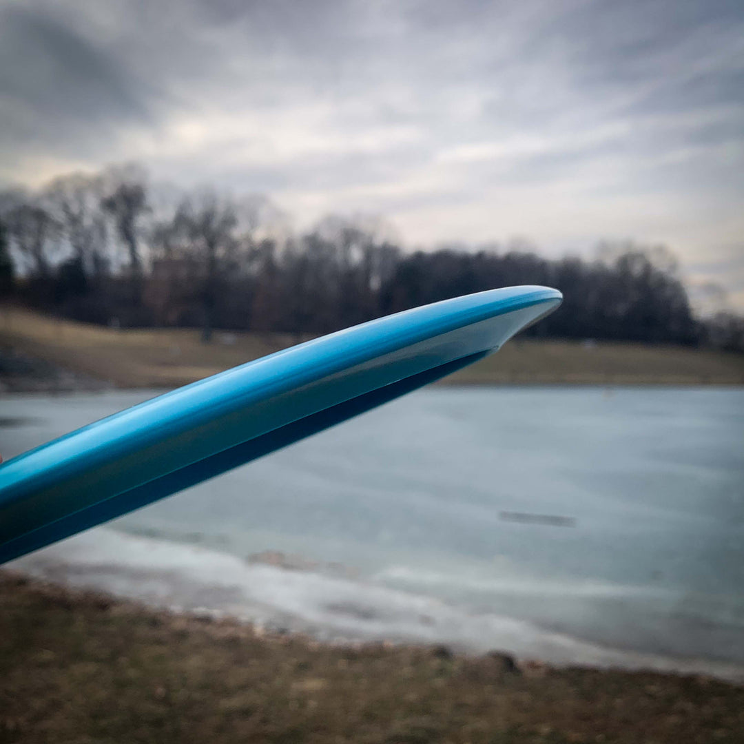 A close-up of the edge of a blue Journey - Diamond long-range distance driver held outdoors near grass and a partially frozen body of water, with trees in the background under a cloudy sky.