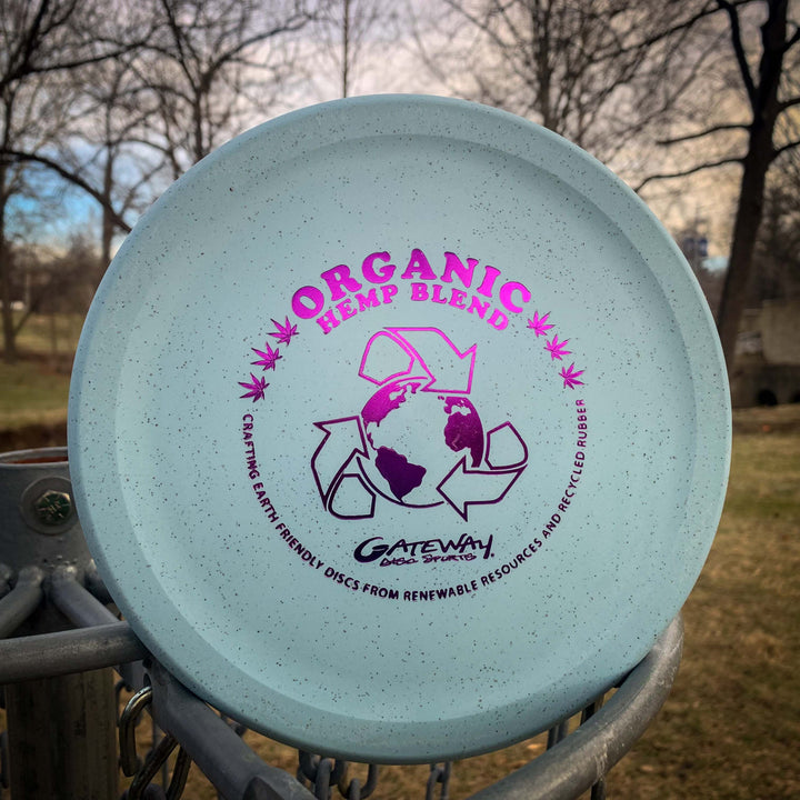 A blue Devilhawk - Suregrip Hemp disc golf putter, labeled "Organic Hemp Blend" with eco-friendly and recycling symbols, is placed on an outdoor disc golf basket. It features an over-stable Thumb Track design and trees in the background.