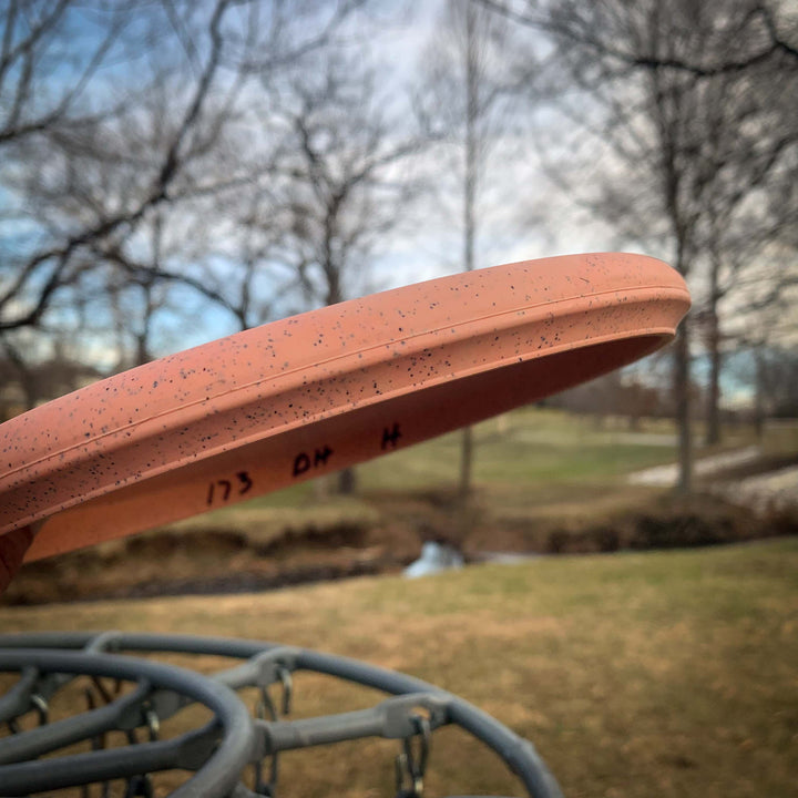 Close-up of an orange Devilhawk - Suregrip Hemp disc golf disc with a Thumb Track design, resting on a metal basket in a park with bare trees and a small creek in the background.