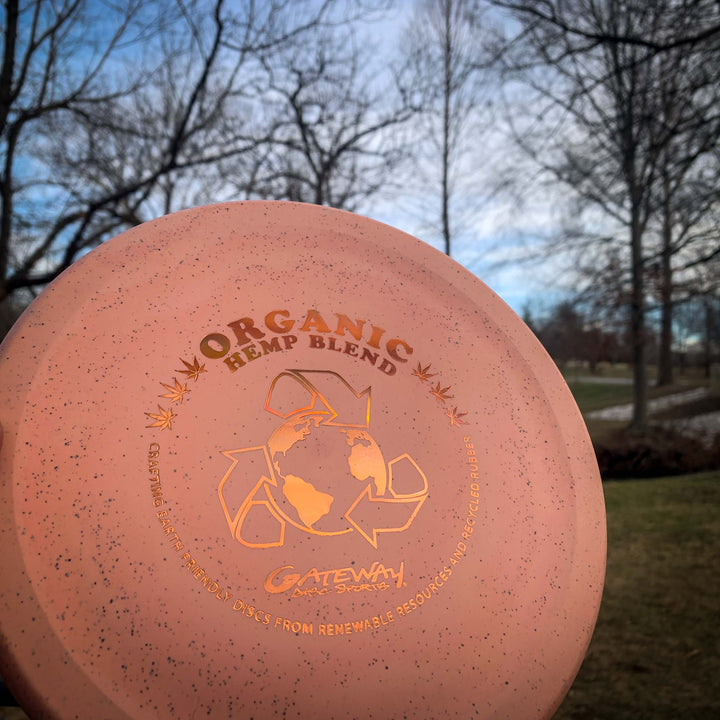 A close-up of a pink Devilhawk - Suregrip Hemp disc golf disc with a Thumb Track design, held outdoors in a park with leafless trees and a cloudy sky in the background.