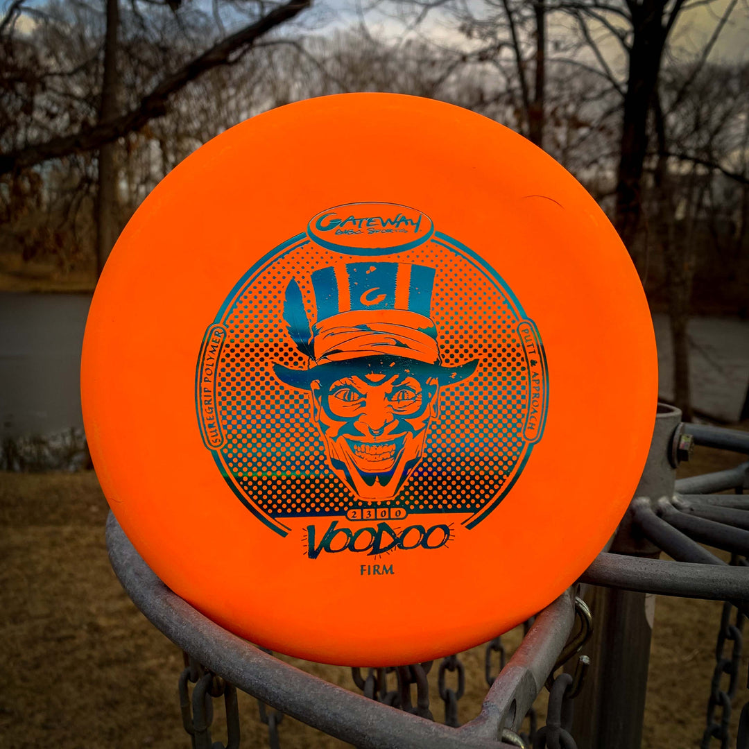 A Voodoo - Suregrip - Firm putter in orange with a blue graphic rests on a disc golf basket, with trees and a pond visible in the background.
