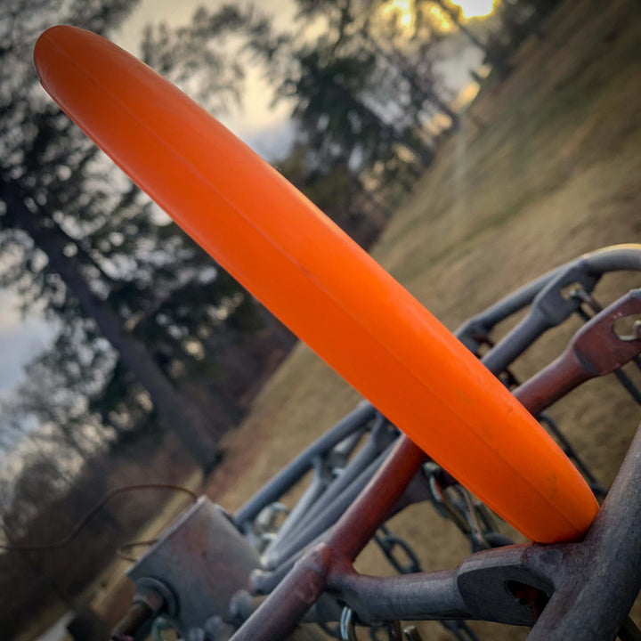 A Voodoo - Suregrip - Firm putter disc golf disc stands upright on a metal basket rim, with trees and a grassy field in the background.