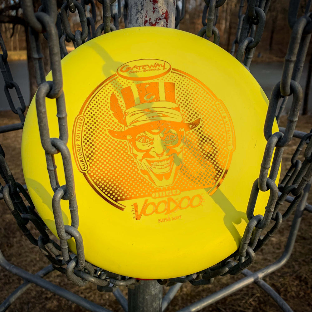 A yellow Voodoo - Suregrip - SS disc golf putter with a grinning top hat character rests in a metal disc golf basket outdoors.
