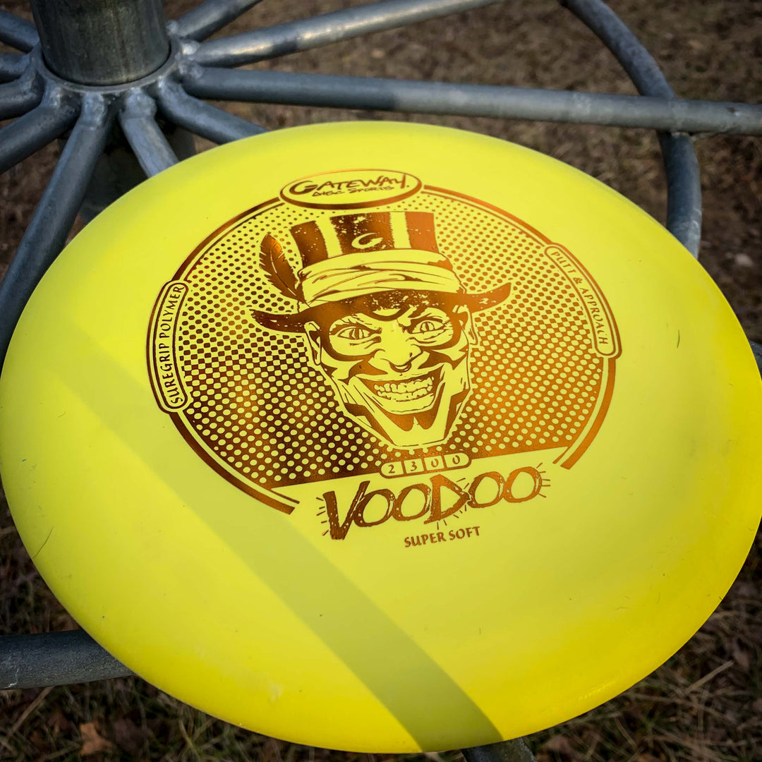 A yellow Voodoo - Suregrip - SS disc golf putter sits on a metal disc golf basket outdoors.