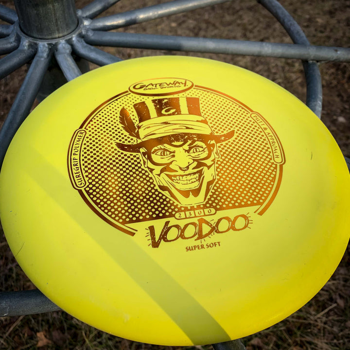 A yellow Voodoo - Suregrip - SS disc golf putter sits on a metal disc golf basket outdoors.