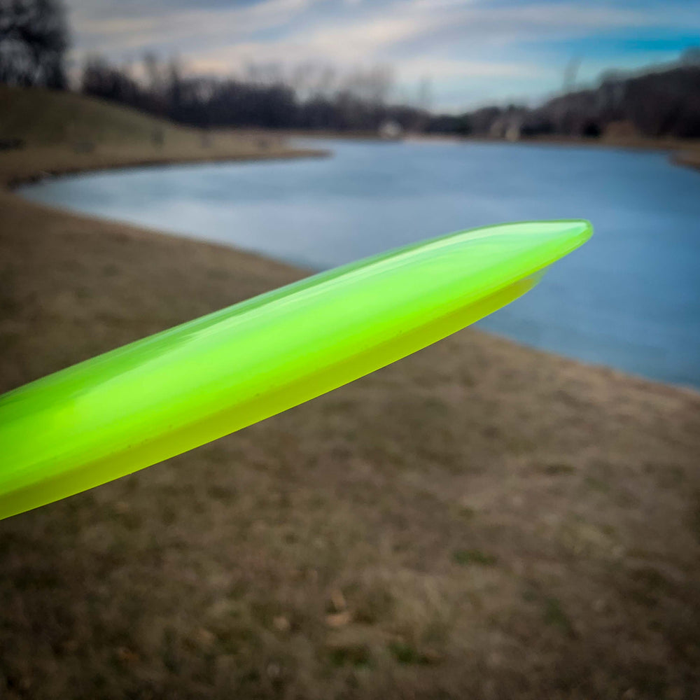 A close-up of the Sabre - Diamond, a green, versatile disc golf disc, held near a grassy pond under a partly cloudy sky.