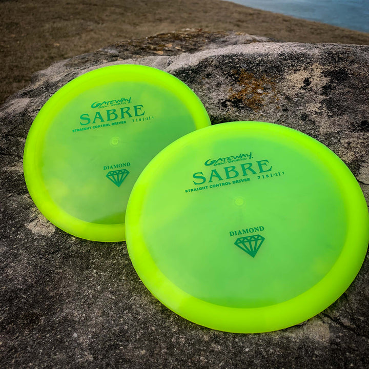 Two yellow-green Sabre - Diamond disc golf discs, known for their straight flight and predictable finish, sit on a large rock outdoors with grass and water in the background.