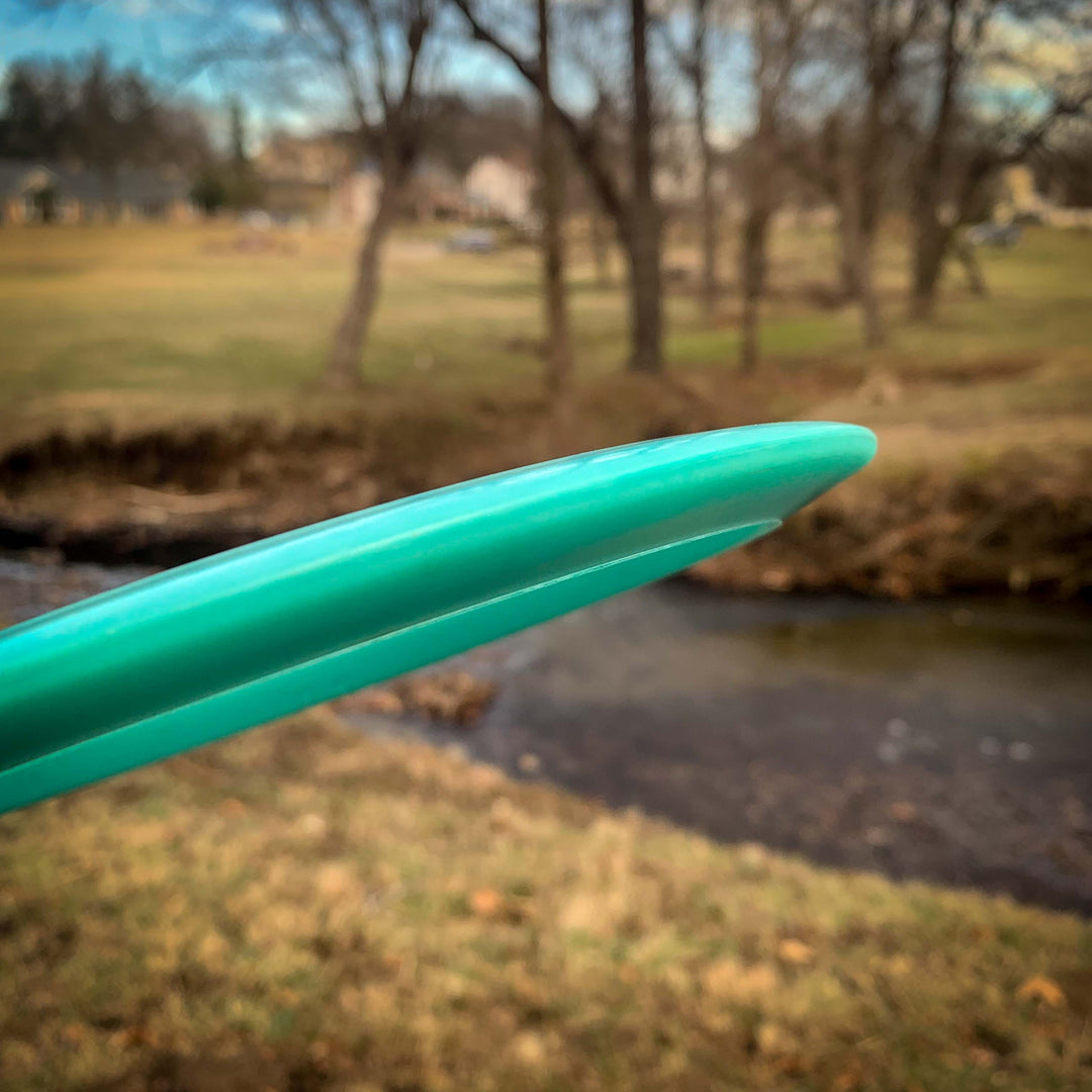 Close-up of a teal Spear - Hyper Diamond (HD) control driver disc golf disc with a comfortable grip. The blurred background features grassy parkland, leafless trees, and a small creek.