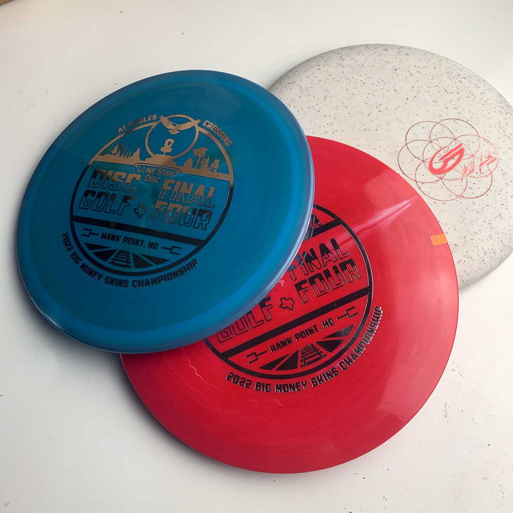 Three disc golf discs are stacked on a white surface: two event-branded blue and red discs from the Big $ Skins Championship at Eagles Crossing, and a white EC Lone Star - Chupacabra disc with a red logo and speckled pattern.
