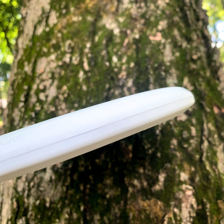 A Magic - Suregrip - SSSS white disc is stuck against the rough, mossy trunk of a tree outdoors.